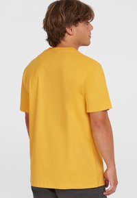 Gul bomulds t-shirt med korte ærmer, rund halsudskæring og en afslappet pasform, vist fra ryggen mod en ensfarvet baggrund.