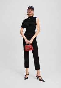 Melna drapēta blūze ar baltām akcentiem, melni piegriezti bikses, sarkana rokassoma ar sudraba logo un melnas siksniņas papēžu kurpes uz vienkārša fona.