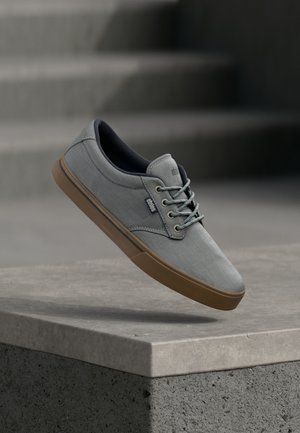 Světlé šedé plátěné tenisky s hnědou gumovou podrážkou, kulatou špičkou a šněrováním. Mají jemné logo a hladký povrch.