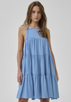 Fiatal nő ujjatlan, világoskék, réteges ruhában, négyzet alakú nyakkivágással és laza fazonnal, egyszínű szürke háttér előtt állva.