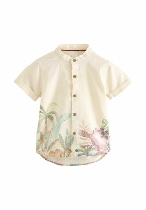 Tout-petit portant un t-shirt beige à manches courtes avec un motif de dinosaure, un short gris et des sandales bleu marine, debout sur un fond orange.