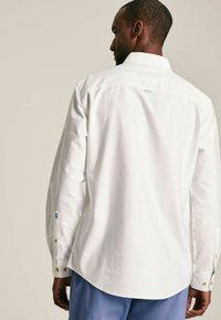 Biała koszula z długim rękawem i kołnierzykiem na guziki, z jedną guzikiem przy mankietach, tylnym karczkiem oraz subtelnym logo blisko kołnierzyka.