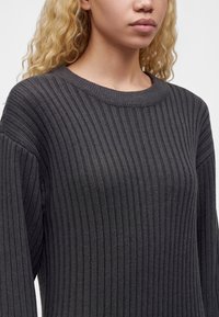Personne aux longs cheveux blonds bouclés portant un pull en maille côtelée gris foncé avec un col rond sur un fond uni.