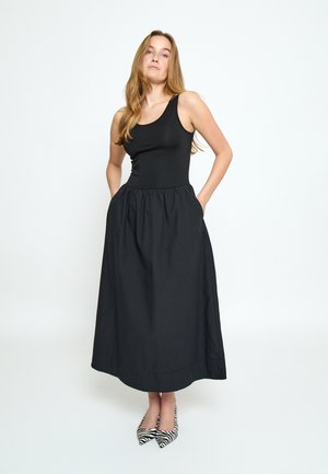 Frau mit langem blondem Haar, die ein ärmelloses schwarzes Kleid mit Taschen und zebra-gestreiften, spitzen Schuhen trägt, vor einem schlichten weißen Hintergrund steht.