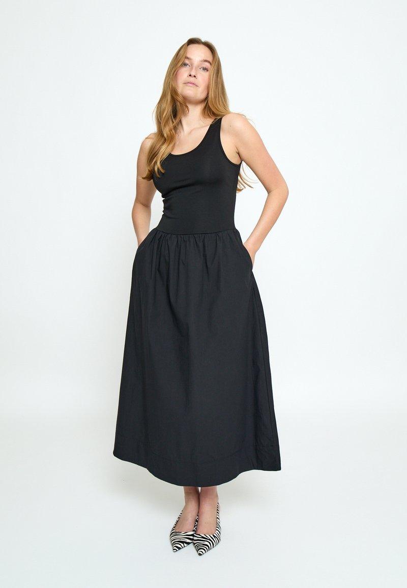 Mujer con cabello largo y rubio, vestida con un vestido negro sin mangas con bolsillos y zapatos puntiagudos con estampado de cebra, de pie frente a un fondo blanco liso.