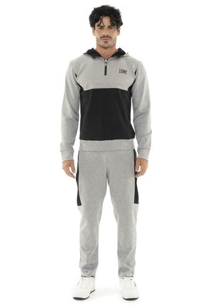 Un hombre luce una sudadera con capucha gris y negra con cremallera hasta el pecho, un pantalón de chándal a juego y zapatillas blancas sobre un fondo blanco.