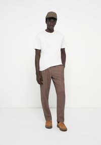 White t-shirt, brown patterned trousers, and beige shoes. The model wears a checkered cap, standing in a neutral background.