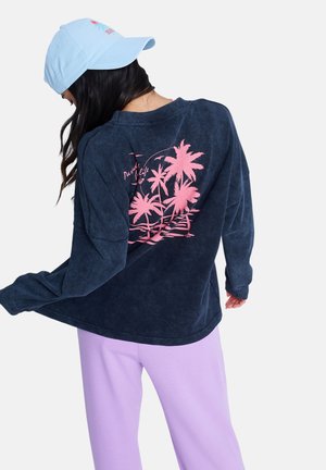 Granatowa bluza z długim rękawem, z różowym nadrukiem palmy na plecach. W zestawie z jasnofioletowymi spodniami i niebieską czapką.