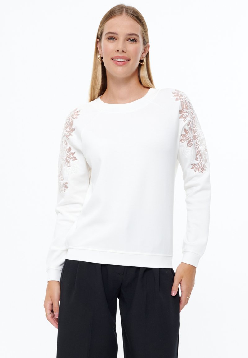 Sudadera blanca con bordados florales en un suave beige en las mangas, cuello redondo y puños acanalados. Combinada con pantalones negros holgados.