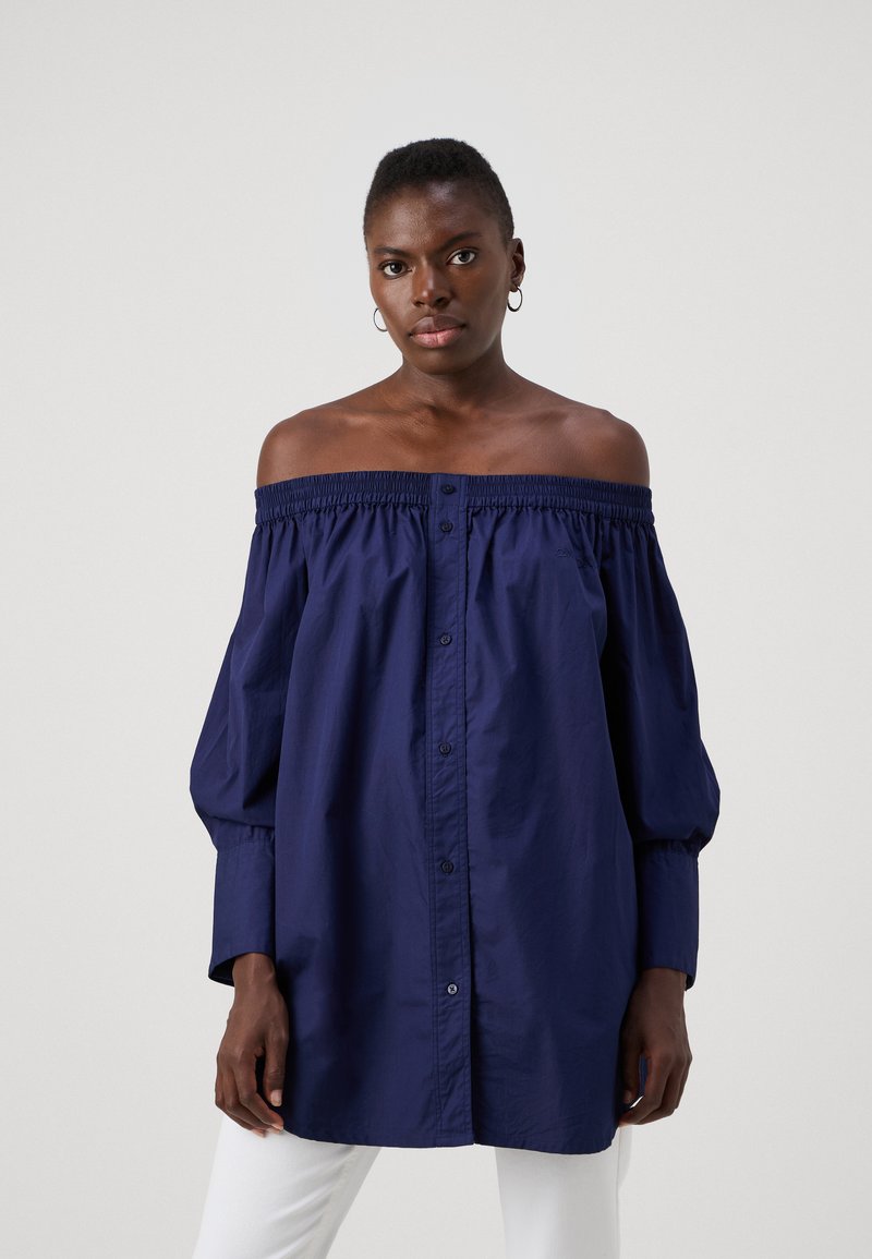 Camicetta navy off-the-shoulder con scollatura elastica, chiusura a bottoni e maniche lunghe a sbuffo. Tessuto morbido con silhouette rilassata.