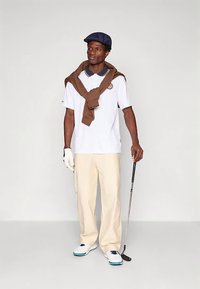 Γκολφ ένδυμα με λευκό πόλο, καφκάι που είναι ριγμένο στους ώμους, κρεμ παντελόνι, μπλε καπέλο και λευκά παπούτσια με μπλε λεπτομέρειες, κρατώντας μια ράβδο.