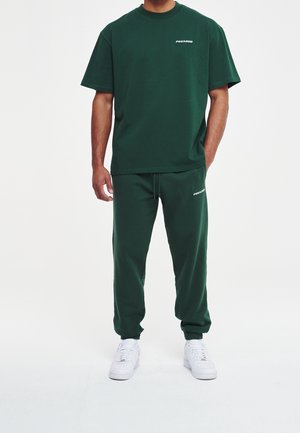 Mężczyzna ubrany w ciemnozieloną koszulkę z krótkim rękawem i dopasowane spodnie dresowe oraz białe trampki na białym tle.