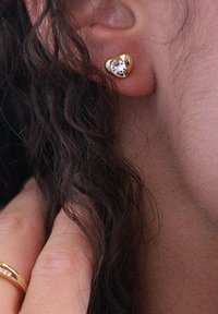 Gli orecchini a perno a forma di cuore in oro presentano una pietra preziosa trasparente al centro, incastonata su una superficie metallica liscia con dettagli lucidati.