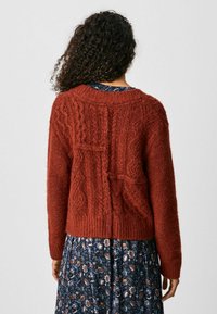 Strickjacke in Rostfarbe mit einem strukturierten Kabeldesign, geripptem Saum und langen Ärmeln. Über einem blumenmusternden Kleid getragen.