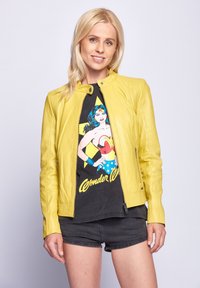 Gelbe Lederjacke mit tailliertem Design, Stehkragen und Reißverschluss auf der Vorderseite. Getragen über einem schwarzen Wonder Woman Grafik-T-Shirt und Shorts.