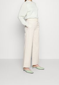 Kvinna iklädd en off-white ribbad tröja som är stoppad i högmidjade vida krämfärgade byxor med bälte, i kombination med ljusgröna slingback kitten heels.