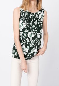 Blouse sans manches à fleurs de couleur vert foncé avec des fleurs blanches et vert clair. Tissu doux, ourlet légèrement courbé, plissage subtil au niveau du col.