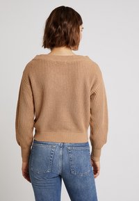 Beige knitted sweater with a loose fit, wide neckline, and ribbed cuffs. Paired with blue high-waisted jeans. Textured fabric detail visible.