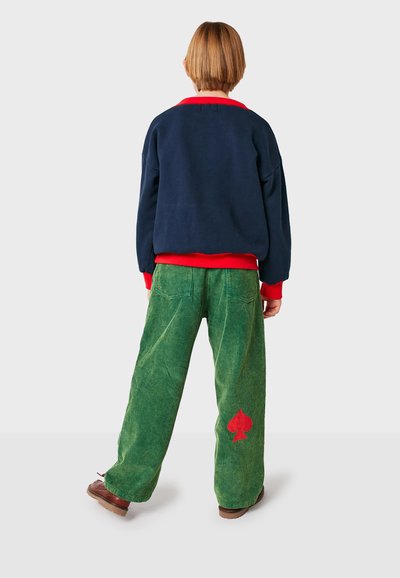 Felpa navy con polsini e colletto rossi, abbinata a pantaloni larghi in corda verde con un'emblema a forma di picche rossa sulla tasca posteriore.