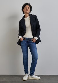 Laivastonsininen villasekoitteinen takki, jossa on leveät kaulukset, kermavärinen karvainen neule, siniset farkut ja valkoiset lenkkarit; rento istuvuus ja rakenteellinen muotoilu.