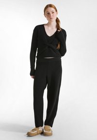 Schwarzer, gerippter Strickpullover mit gedrehtem Vorderteil und langen Ärmeln, kombiniert mit schwarzen weit geschnittenen Hosen und beigefarbenen Slippern mit Furr-Akzenten.