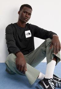 Schwarzes Langarmhemd mit einem weißen Logo auf der Brust, kombiniert mit hellgrünen Hosen. Die Person sitzt auf einem blauen Teppich und trägt schwarze Schuhe.