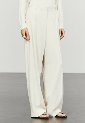 Mannequin portant un pantalon large, ample, taille haute, couleur crème, associé à des sandales noires ouvertes à l'avant sur un sol gris.