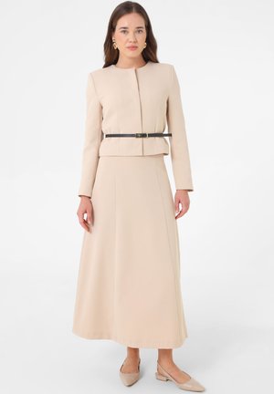 Donna con capelli castani lunghi che indossa una giacca beige a maniche lunghe, gonna beige lunga fino alla caviglia, cintura nera e scarpe beige con punta, in piedi davanti a uno sfondo bianco.