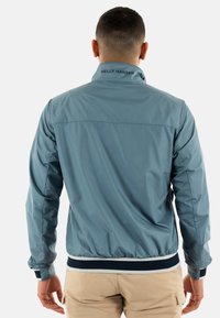 Giacca leggera blu con colletto alzato, polsini elasticizzati e orlo a righe bianche e blu navy. Presenta un logo sul colletto posteriore.