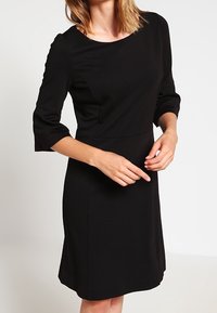 Czarna sukienka do kolan z rękawami o długości trzech kwart, gładki materiał, okrągły dekolt i dopasowany gorset, rozszerzająca się na spódnicy.