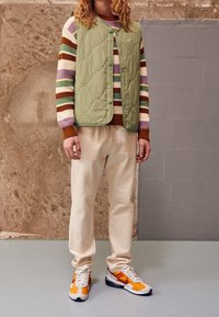 Person som bär beige byxor, orangea och vita sneakers, en grön vadderad väst och en randig tröja med lila, bruna och gröna band.