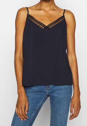 Femme portant un haut noir sans manches avec une bordure en dentelle au niveau du décolleté et un jean en denim bleu, debout devant un fond uni.