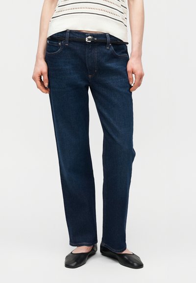 Άτομο που φοράει σκούρα μπλε τζιν με ίσιο κόψιμο, μαύρη ζώνη, λευκό αμάνικο πλεκτό τοπ και μαύρες μπαλαρίνες σε λευκό φόντο.