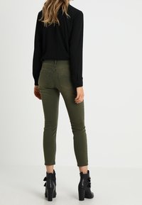 Pantalones ajustados de color verde oliva con una textura suave, combinados con una blusa negra de manga larga y botas de tobillo negras con tacón y detalles de lazo.