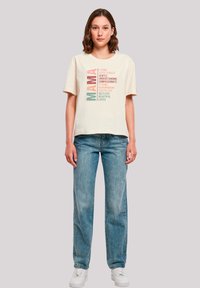 Beige cotton t-shirt with short sleeves, featuring bold, colorful text on the front that lists positive attributes. Worn with light blue jeans.