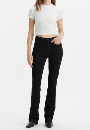 Kobieta ubrana w biały krótki top z krótkim rękawem, czarne jeansy z wysokim stanem i prostymi nogawkami oraz czarne spiczaste buty na jednolitym tle.