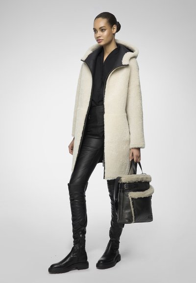 Donna che indossa un cappotto di shearling beige, pantaloni di pelle nera, stivali neri e porta una borsa nera con rifiniture in shearling, su uno sfondo semplice.