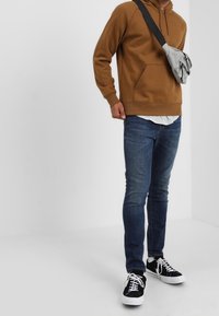 Person som bär en brun huvtröja, blå jeans, svarta sneakers och en grå axelväska mot en enkel vit bakgrund.