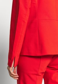 Gros plan sur la main d'une personne reposant à côté de la hanche, portant un costume rouge vif sur mesure avec une légère fente à la manche.