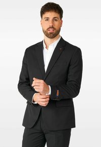 Chaqueta de traje negra hecha de un material suave. Presenta un único bolsillo en el pecho, texto naranja "DAILY" en la manga y una camisa blanca con cuello debajo.