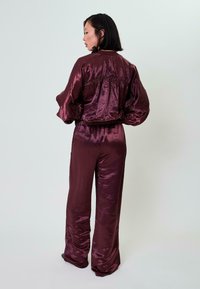 Burgundi satino kostiumas, turintis susiraukiantį, elastingą liemenį, didelėmis rankovėmis ir atitinkamas plačias kelnes su užpakaline kišene.