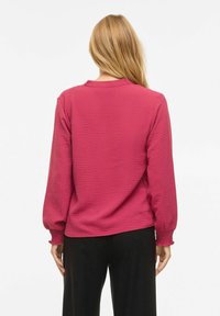 Femme aux longs cheveux blonds portant un haut rose à manches longues texturé et un pantalon noir, debout de dos sur un fond blanc.