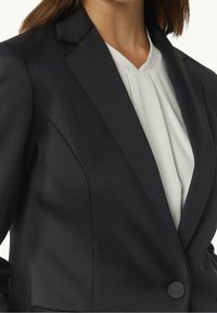 Blazer noir à texture lisse, avec des revers crantés et une fermeture à un bouton. En dessous, un chemisier plissé de couleur claire est visible.