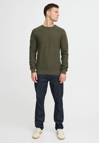 Olivgrünes Langarm-Rippstrickoberteil, kombiniert mit dunkelblauen Jeans und weißen Sneakers mit beigen Akzenten. Schlichtes, tailliertes Design.