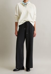 Modèle portant une chemise polo blanche à manches longues, un pantalon noir ample avec cordon de serrage, un foulard de cou bleu clair et des mocassins noirs sur fond uni.