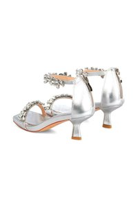 Sandalias metálicas plateadas con tacones pequeños, que presentan un diseño de punta abierta y correas adornadas con cristales transparentes. Cierre de cremallera en la parte trasera.