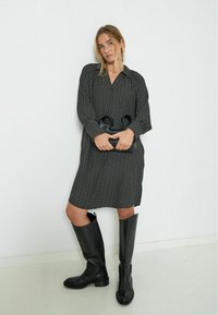 Schwarzes und weißes gemustertes Kleid mit langen Ärmeln, Knopfverschlusskragen und geradem Schnitt. Kombiniert mit hohen schwarzen Lederstiefeln und einer schwarzen Tasche.