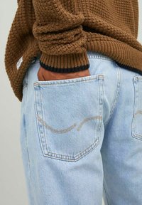 Un jean en denim bleu clair avec des coutures visibles sur les poches, associé à un pull en maille brun texturé avec un poignet côtelé bleu marine.
