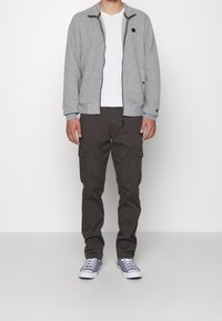 Giacca imbottita grigia con zip, dettagli neri, maglietta bianca, pantaloni cargo scuri e sneakers grigie. Caratterizzata da tasche frontali e polsini a costine.