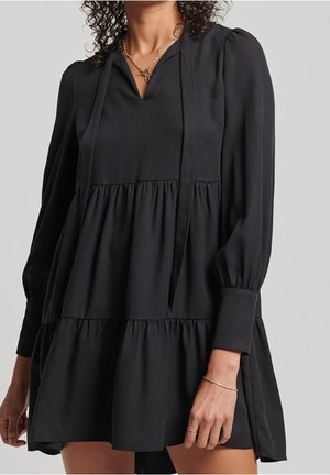 Kobieta w czarnej mini sukience z długim rękawem, o warstwowym kroju, z dekoltem w kształcie litery V i wiązaniem na szyi, dopełniona złotym naszyjnikiem i bransoletką.